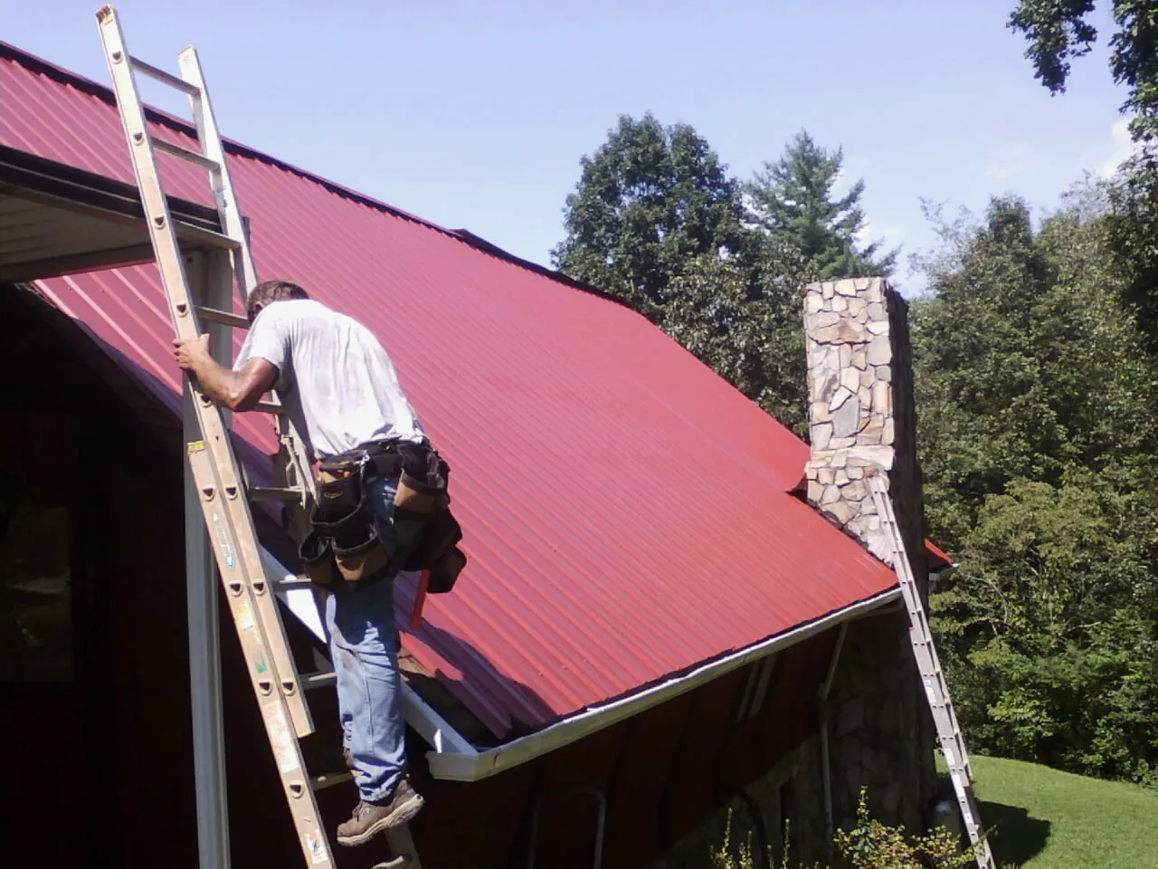 Roofer on a ladder beside a red metal roof and stone chimney — Soffit & Fascia Repair by Roofing Brothers in Maple Lake